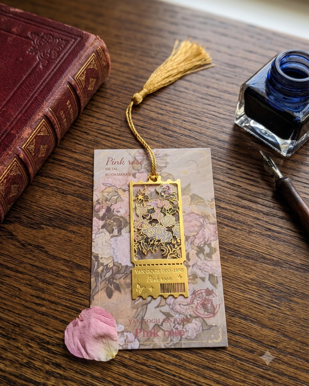 A gold-toned metal bookmark with a tassel and floral cutout design, displayed on a decorative backing card on a wooden table. A pink rose petal, an antique-style red book, a blue ink bottle, and a fountain pen are arranged around it.