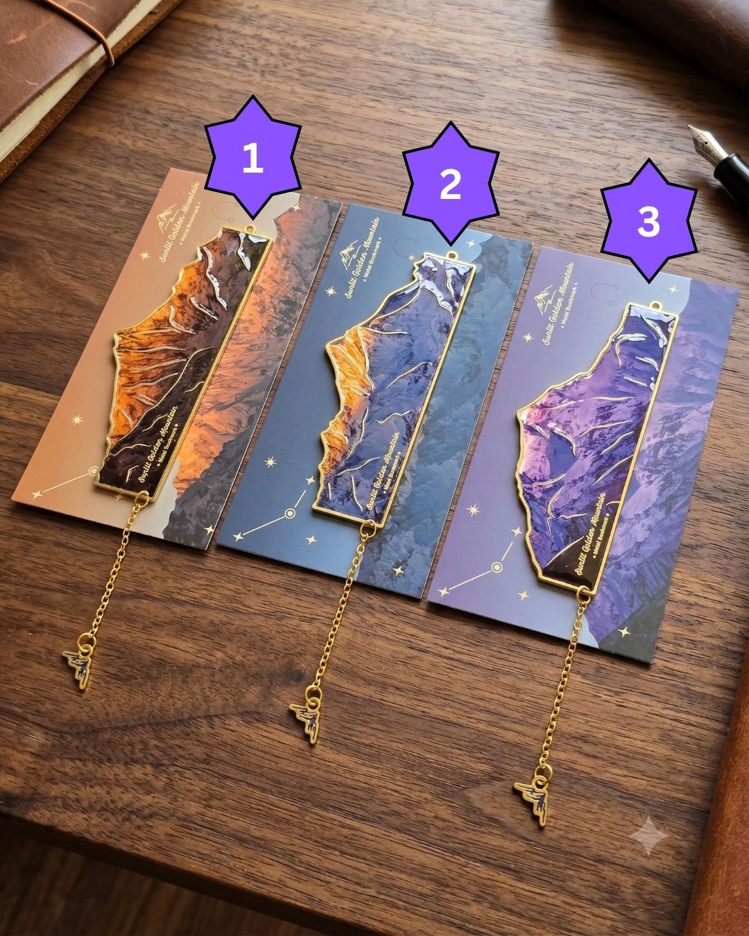 Three bookmarks featuring mountain landscapes in shades of orange, blue, and purple, each with a gold chain and charm.
