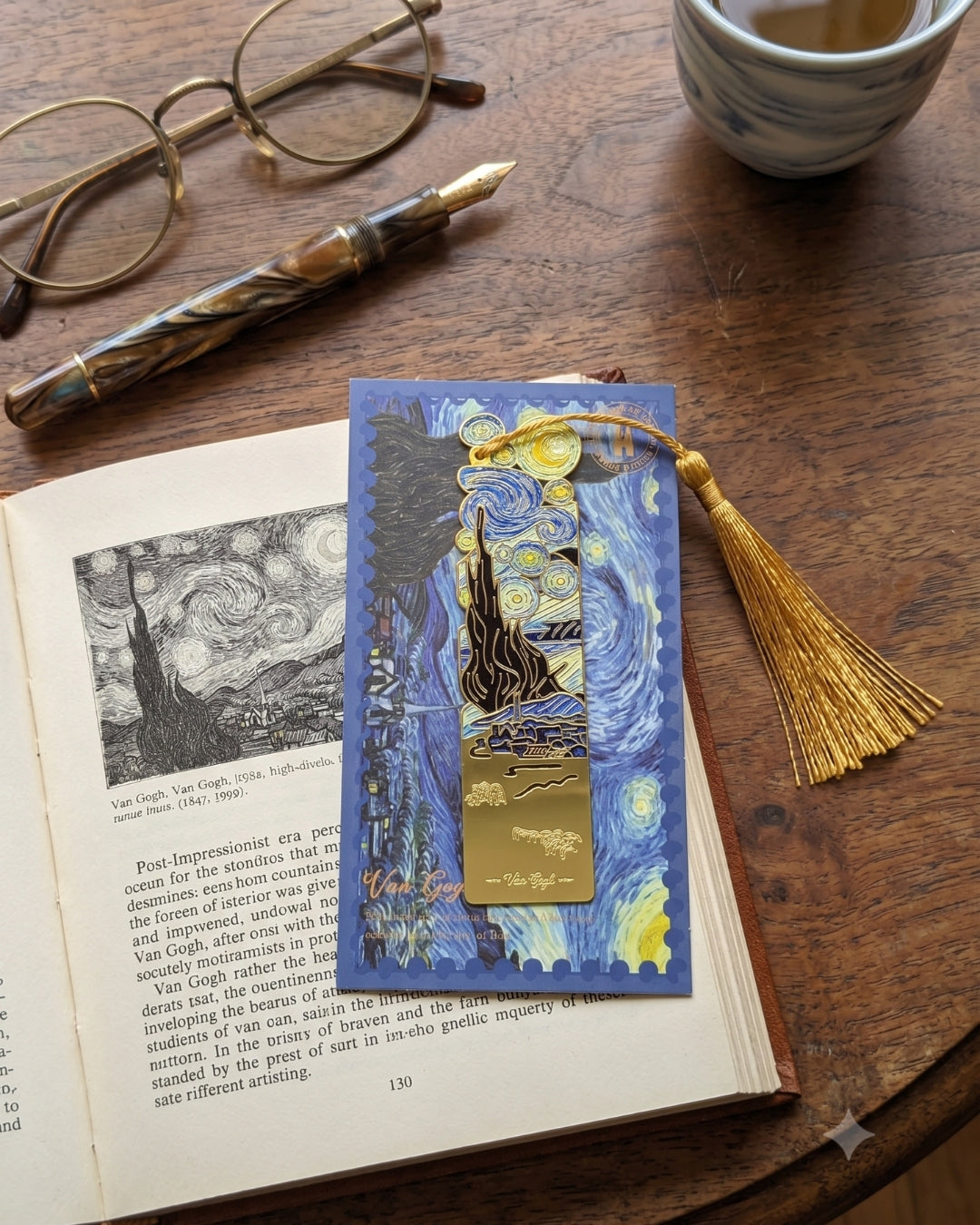 A bookmark featuring Van Gogh's Starry Night design rests on an open book next to a fountain pen and glasses on a wooden table.