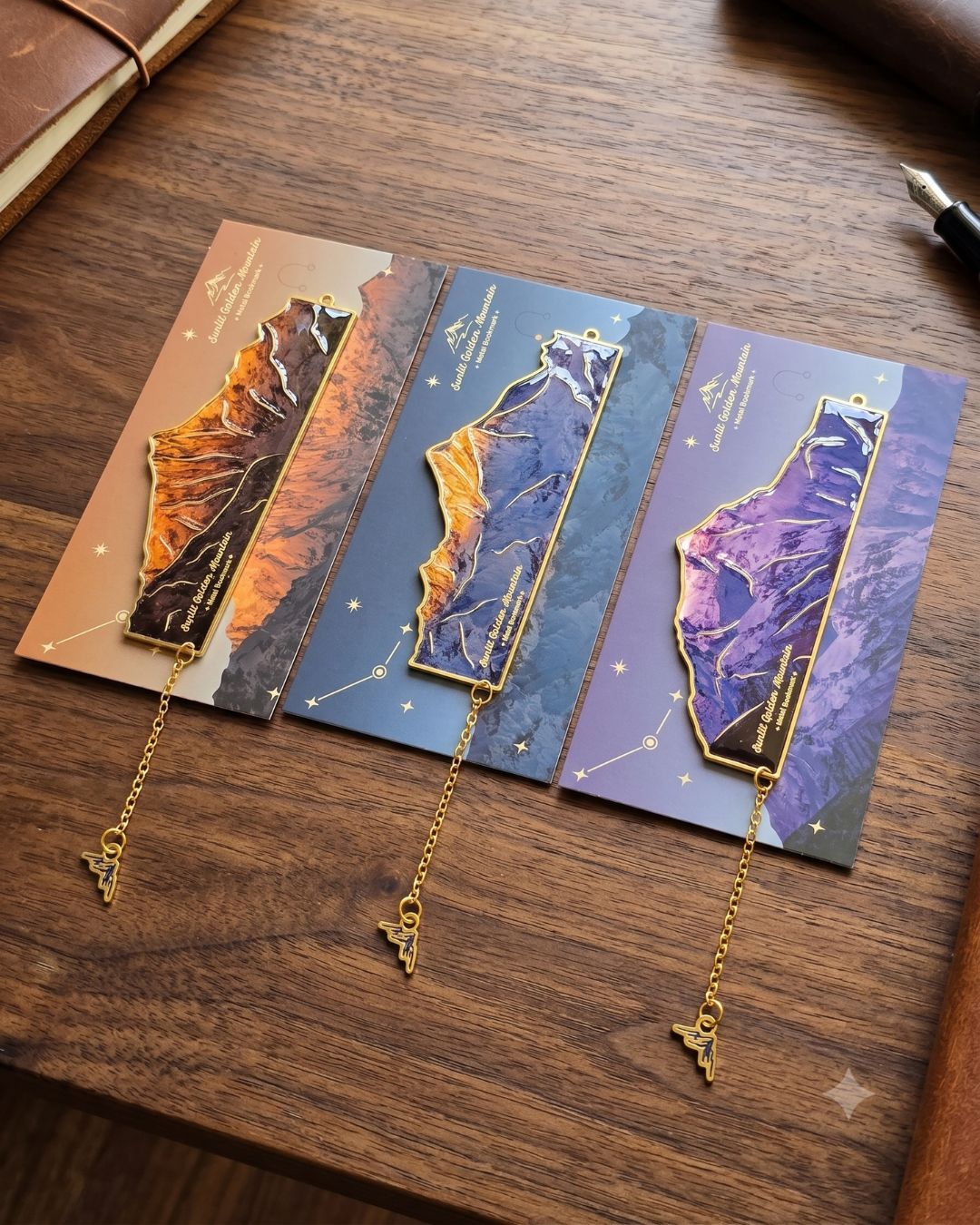 Three landscape-themed bookmarks with mountain designs in shades of orange, blue, and purple, each with a gold chain and charm.