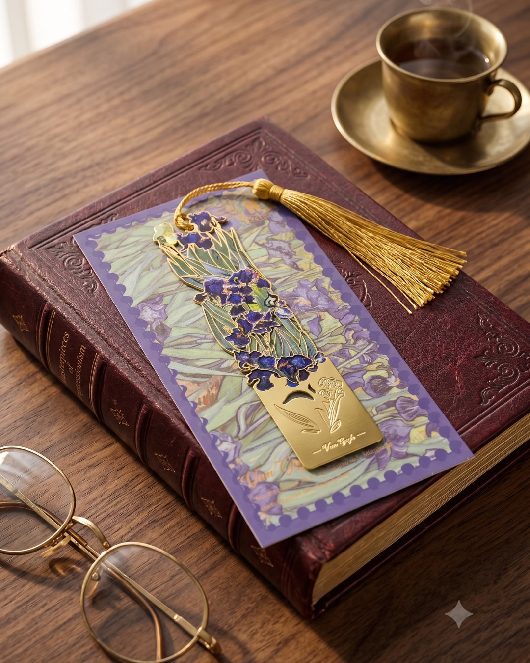 A golden bookmark with a purple floral design and a gold tassel rests on a closed book next to a pair of glasses and a cup of tea.