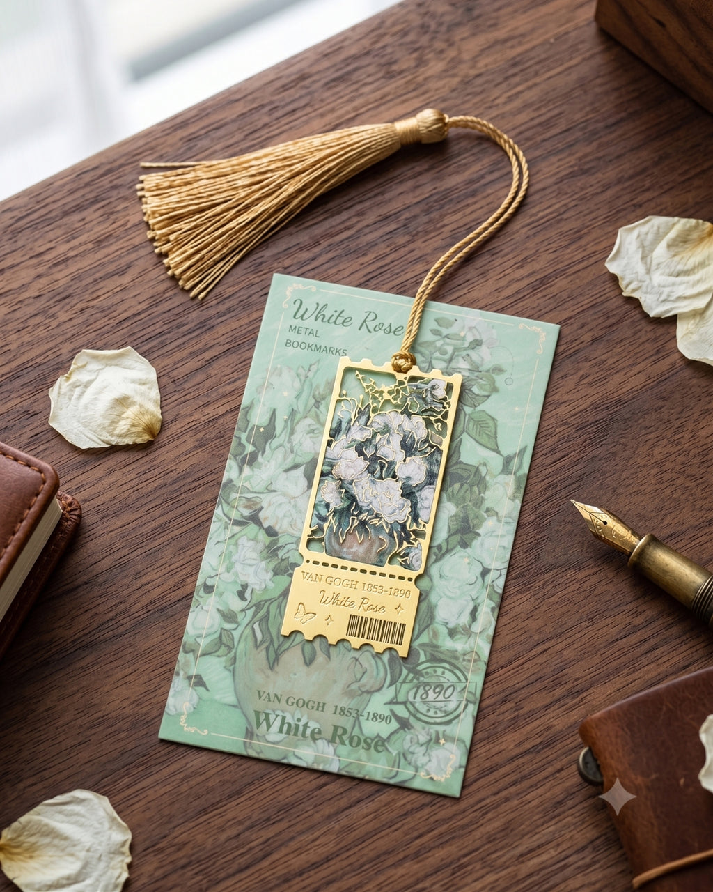 A decorative metal bookmark with a gold tassel and a rectangular floral pendant, displayed on a wooden desk with dried petals, a pen, and leather accessories. The card reads 'White Rose Metal Bookmark'.