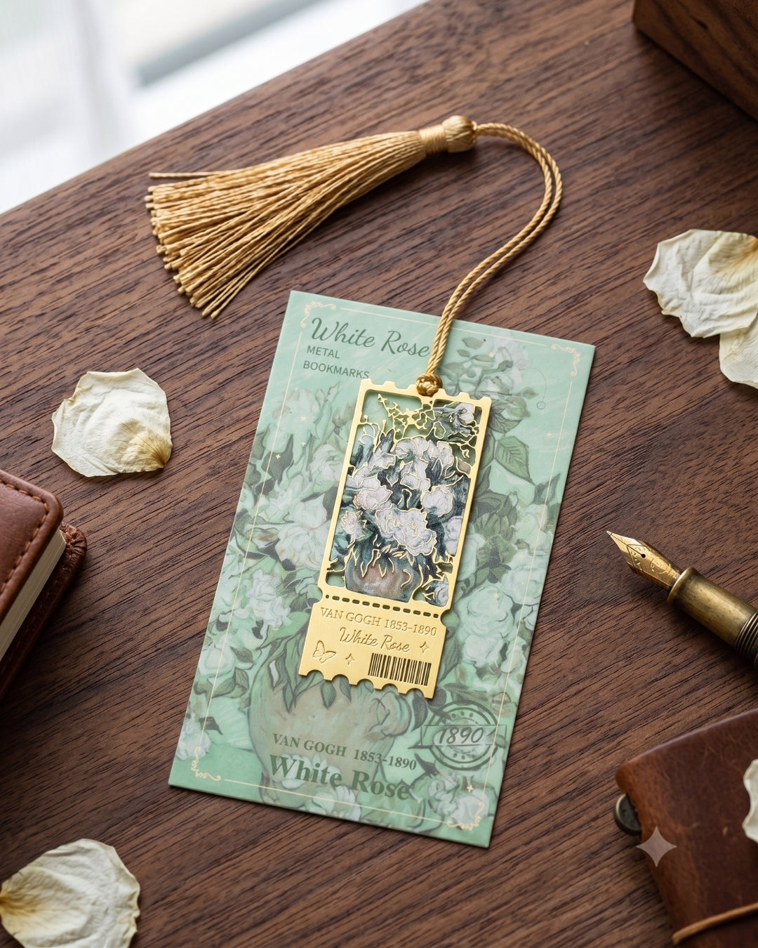 A decorative metal bookmark with a gold tassel and a rectangular floral pendant, displayed on a wooden desk with dried petals, a pen, and leather accessories. The card reads 'White Rose Metal Bookmark'.