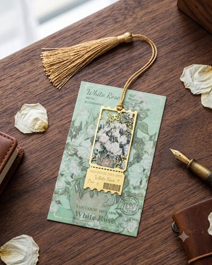 A decorative metal bookmark with a gold tassel and a rectangular floral pendant, displayed on a wooden desk with dried petals, a pen, and leather accessories. The card reads 'White Rose Metal Bookmark'.