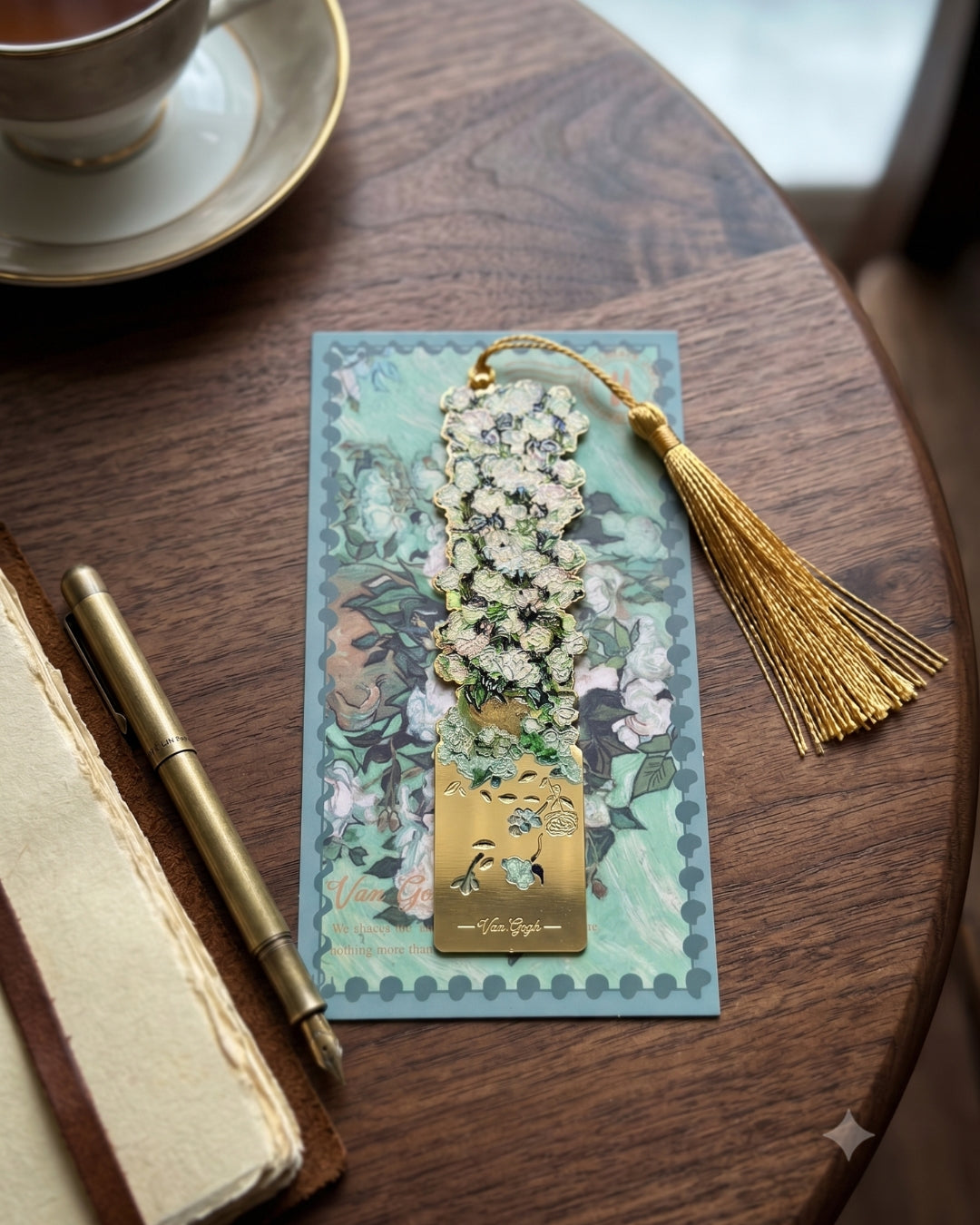 A decorative bookmark with a floral design and a gold tassel rests on a postcard with a floral pattern. A pen and a cup of tea are visible nearby.