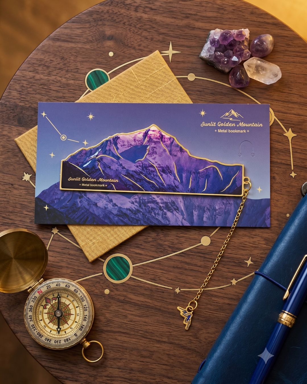 A flat lay of celestial-themed stationery, featuring a mountain landscape postcard, a compass, a blue notebook, a pen, and amethyst crystals on a wooden table with gold constellation designs resembling a night sky.