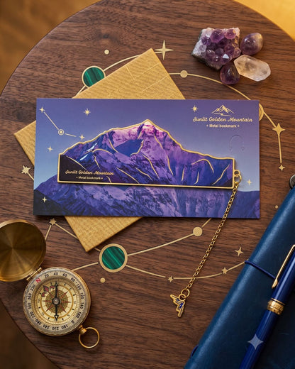A flat lay of celestial-themed stationery, featuring a mountain landscape postcard, a compass, a blue notebook, a pen, and amethyst crystals on a wooden table with gold constellation designs resembling a night sky.