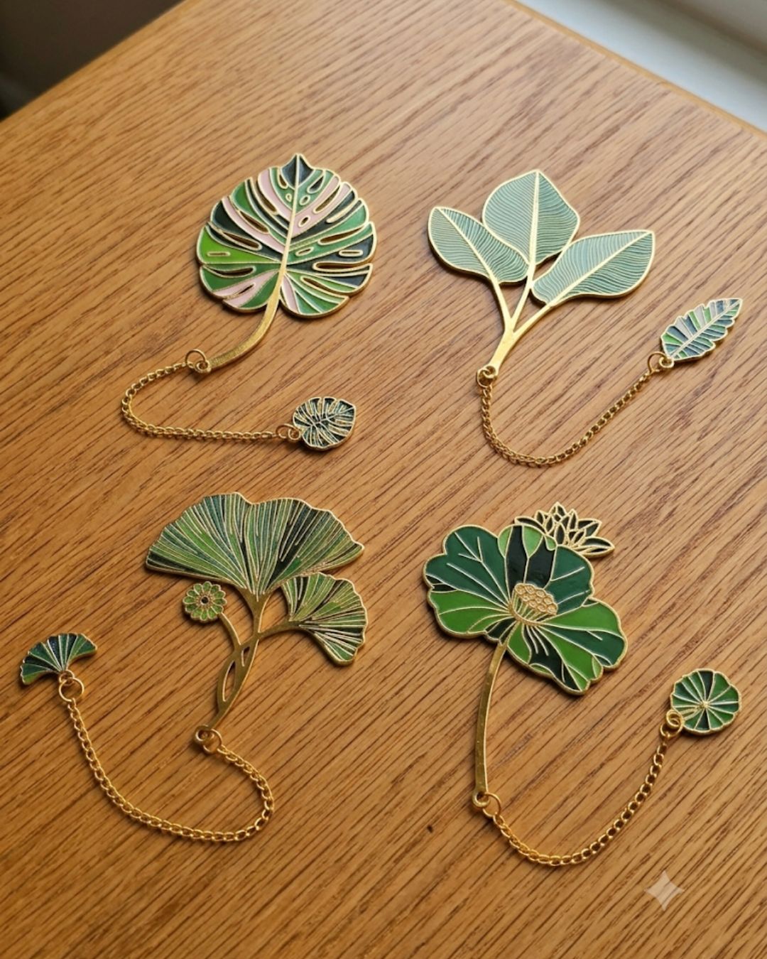 Four decorative enamel bookmarks shaped like leaves and flowers with gold chains on a wooden surface.