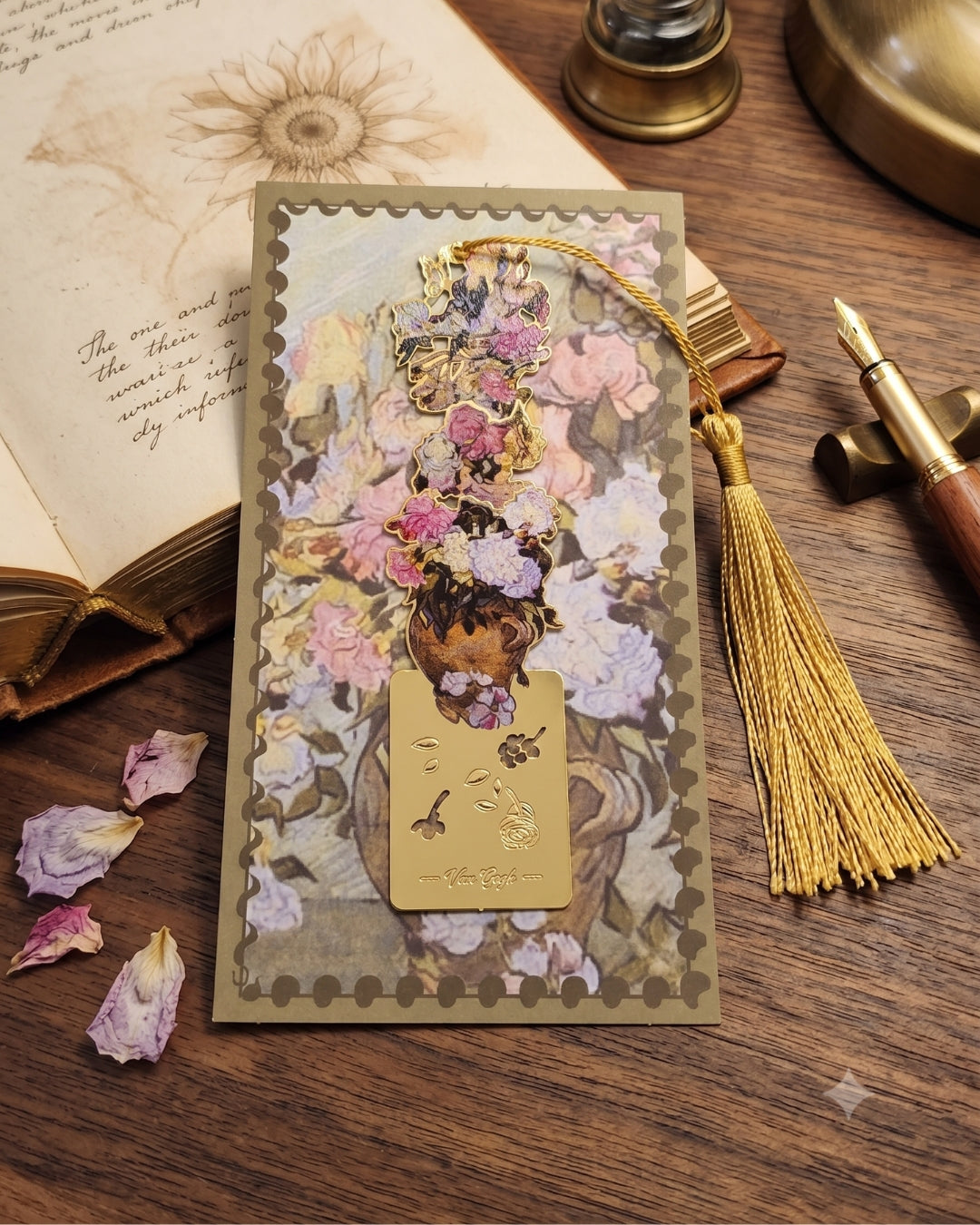 A bookmark featuring a floral arrangement and a gold tassel, placed on a wooden desk next to an open book and dried flower petals.