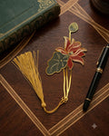 A gold bookmark shaped like a lotus flower with red petals and a green leaf, attached to a gold tassel, rests next to a green book and a black pen on a wooden desk.