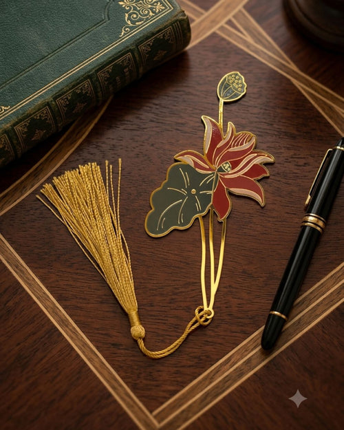 A gold bookmark shaped like a lotus flower with red petals and a green leaf, attached to a gold tassel, rests next to a green book and a black pen on a wooden desk.
