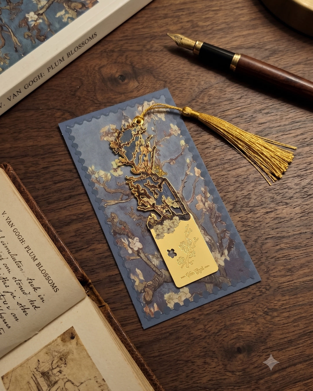 A gold metal bookmark with a tassel rests on a blue bookmark featuring Van Gogh's Plum Blossoms, next to an open book and a fountain pen.