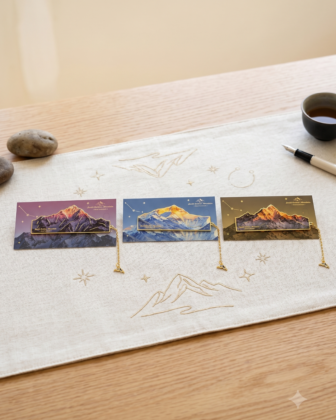 Three bookmarks featuring mountain landscapes in different lighting conditions, displayed on a textured runner with embroidered mountain motifs.