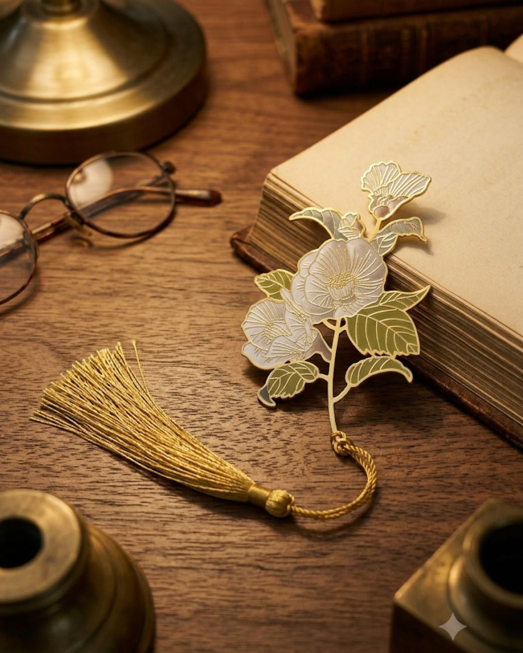 A gold floral bookmark with a tassel rests on an open book next to reading glasses on a wooden desk.