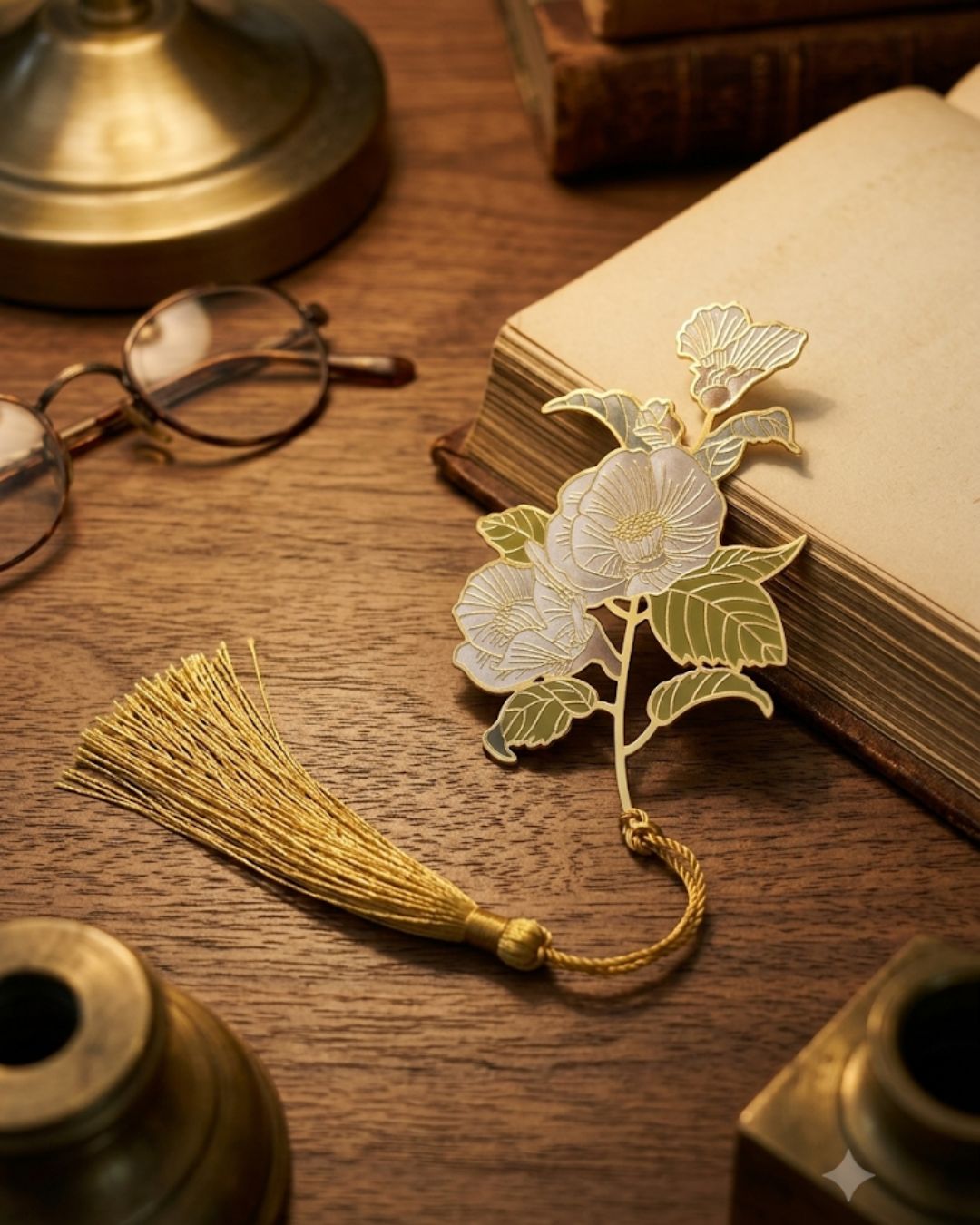 A gold floral bookmark with a tassel rests on an open book next to reading glasses on a wooden desk.