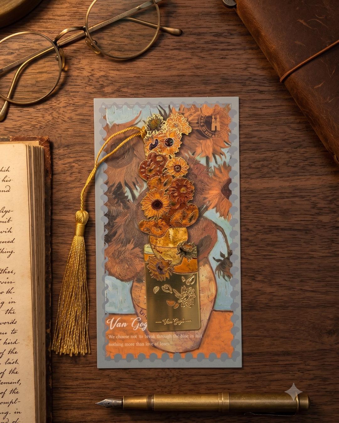 A decorative bookmark featuring Vincent van Gogh's Sunflowers, with a golden tassel and gold foil accents, resting on a wooden desk next to an open book and glasses.