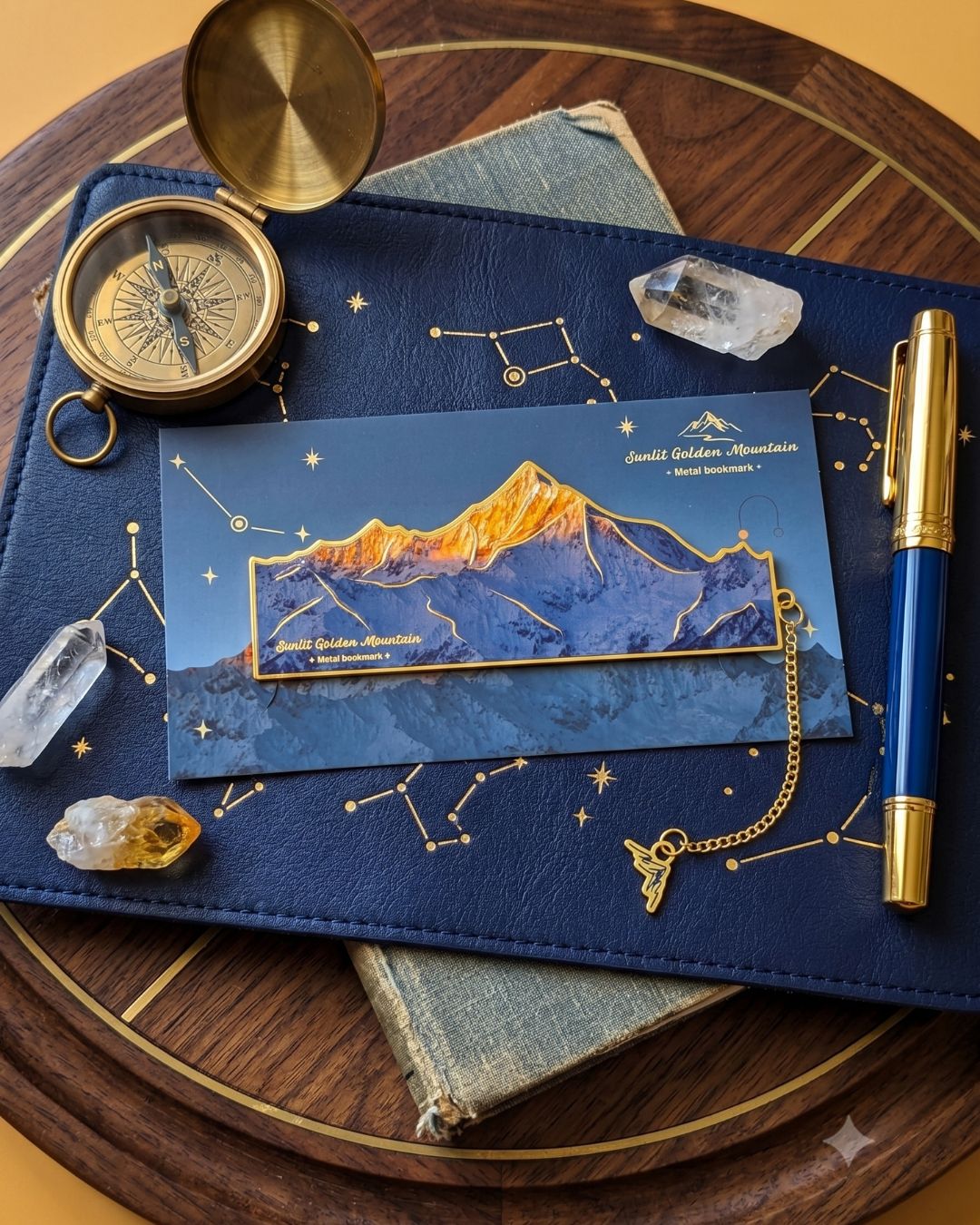 A dark blue notebook with constellations printed on it, featuring a bookmark of a golden mountain range. A vintage compass, a blue and gold pen, crystals, and an old book are also present.