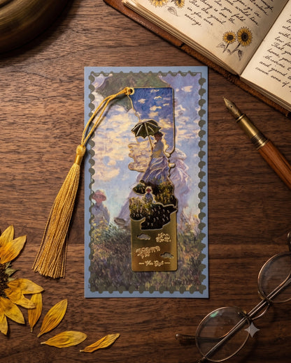 A bookmark featuring a scene from Claude Monet's painting "Woman with a Parasol - Madame Monet and Her Son" is placed on a blue postcard-like background with a gold tassel. The scene depicts a woman holding a parasol, with another figure in the background. The bookmark and postcard are on a wooden surface next to an open book with sunflowers, a fountain pen, and glasses.