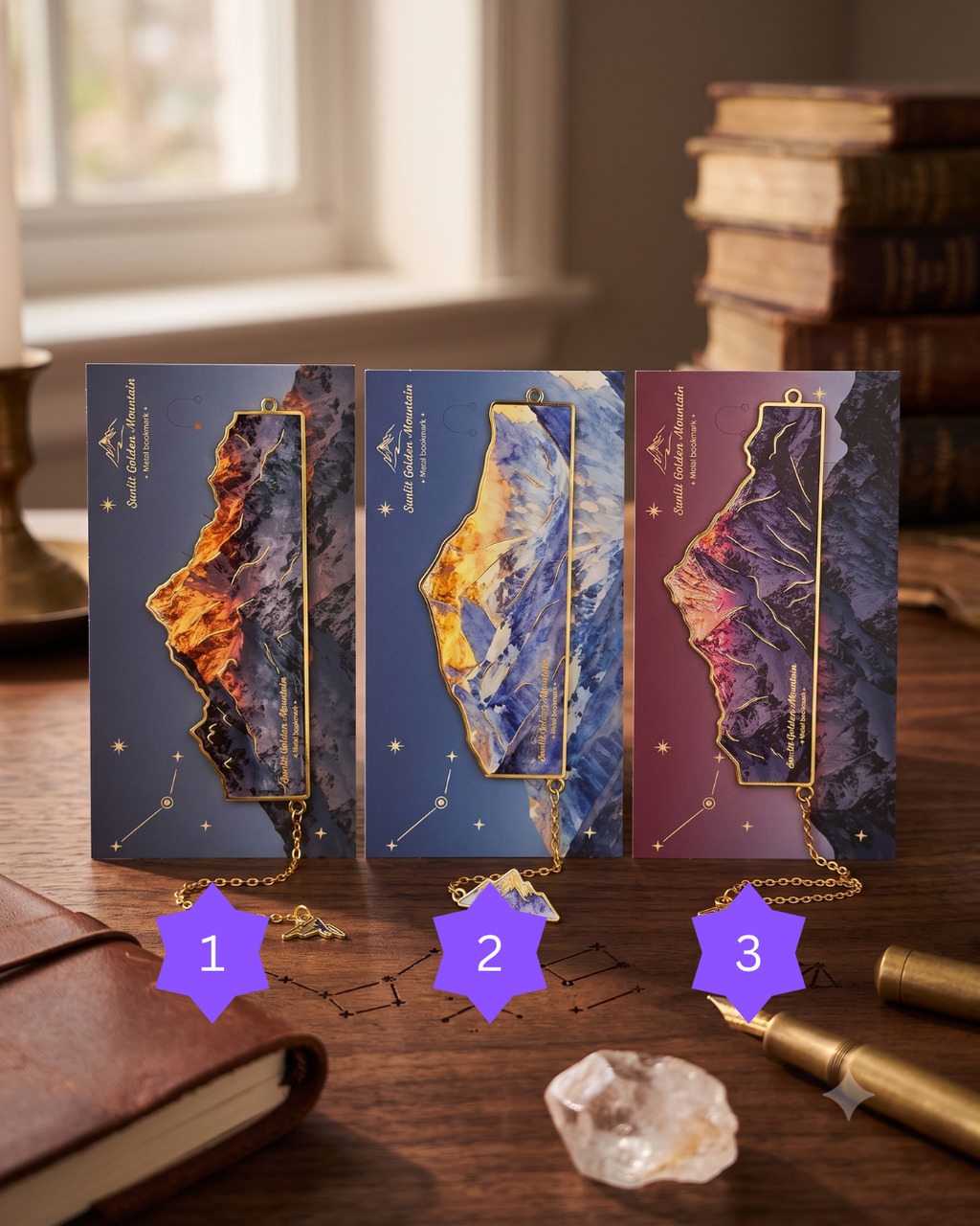 Three ornate bookmarks with mountain and celestial designs, featuring gold accents and small gemstone charms, rest on a wooden desk near a leather-bound journal and a stack of books.