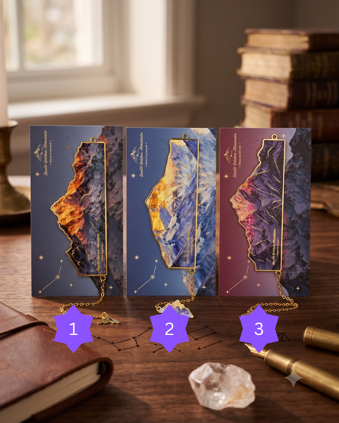 Three ornate bookmarks with mountain and celestial designs, featuring gold accents and small gemstone charms, rest on a wooden desk near a leather-bound journal and a stack of books.