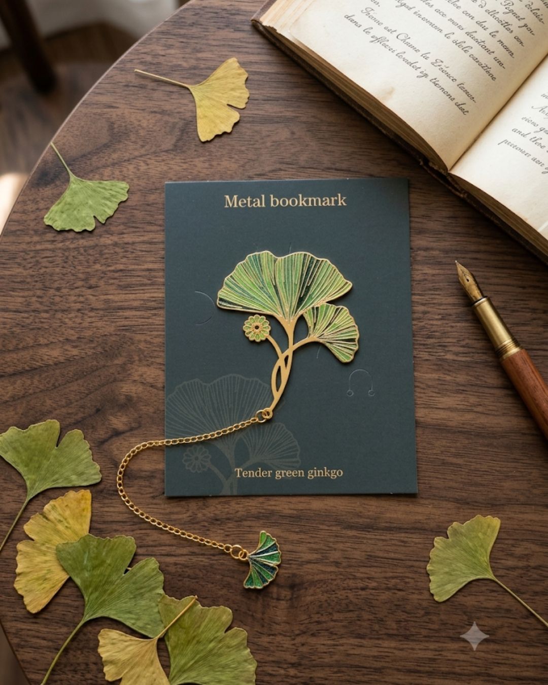 A metal bookmark shaped like a ginkgo branch with green leaves is displayed on a dark surface next to an open book and a fountain pen. Several real ginkgo leaves are scattered around the bookmark.