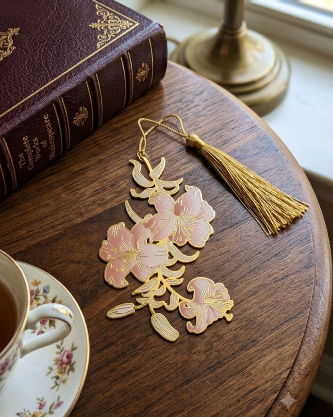 A gold metal bookmark shaped like a branch with pink lily flowers and a gold tassel rests on a wooden table next to a book and a teacup.