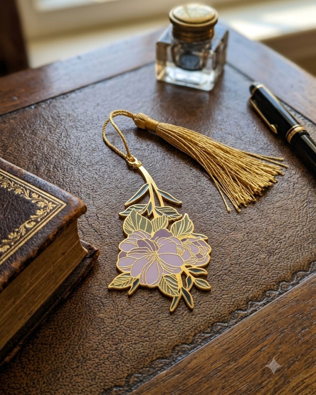 A gold tassel bookmark with a floral design featuring purple flowers and green leaves rests on a leather-bound book next to an inkwell and pen.