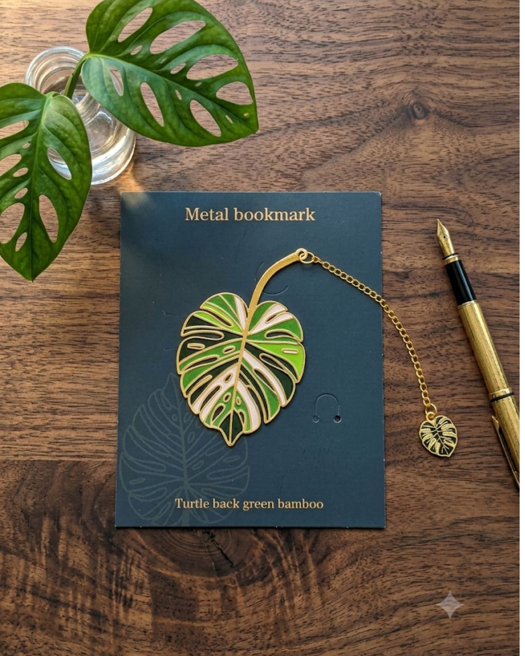 A metal bookmark shaped like a monstera leaf in shades of green, white, and black, attached to a gold chain with a smaller leaf charm. It is presented on a dark green card with text that reads "Metal bookmark" and "Turtle back green bamboo." A glass of water with monstera leaves and a gold pen are also visible on a wooden surface.