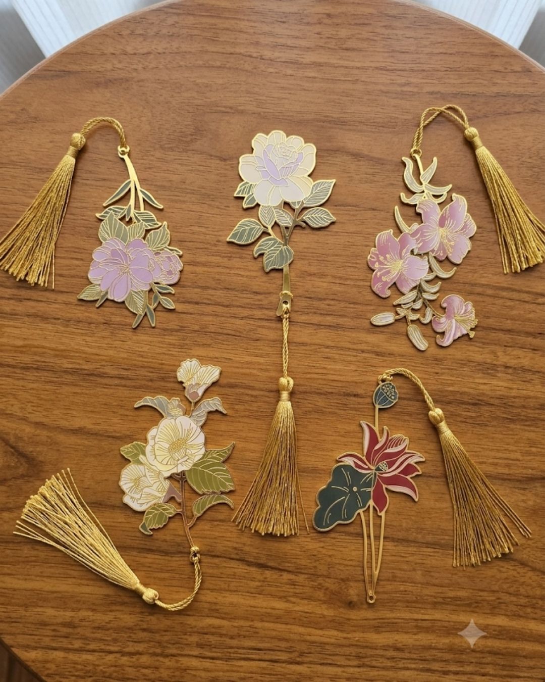 A collection of five decorative bookmarks, each featuring a floral design with gold tassels, laid out on a wooden surface.